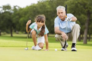 Senior adult Caucasian man is golf pro at country club. He is giving private golf lesson to elementary age Hispanic little girl. He is teaching student how to line up golf ball with putter on green course.